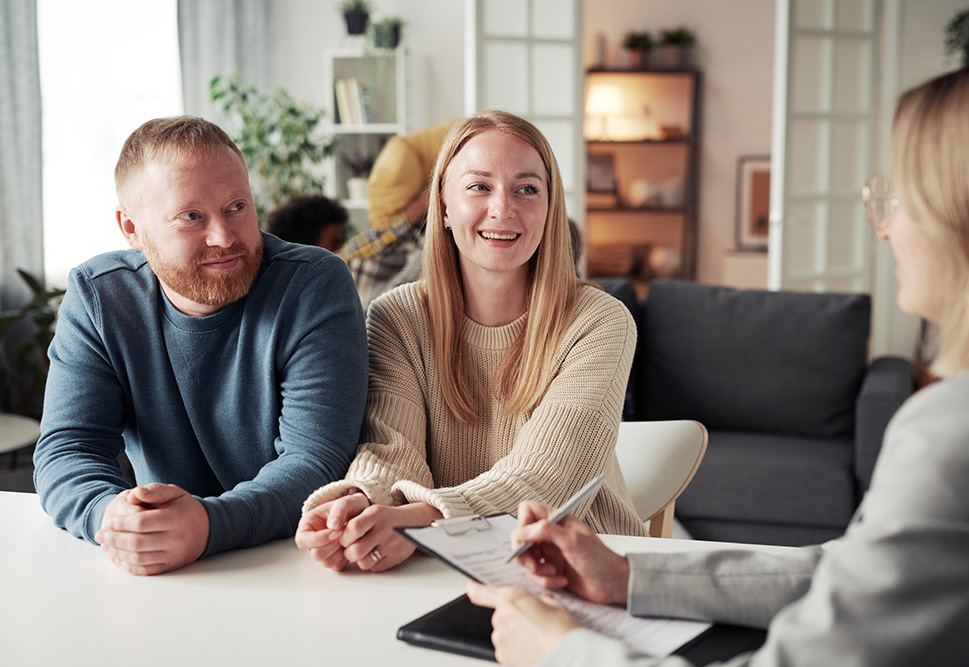 Man and a woman at a table in discussion with a woman writing on a clipboard.